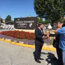 A news reporter films his report in from of a sign welcoming visitors to the CUA campus.