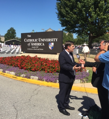 A news reporter films his report in from of a sign welcoming visitors to the CUA campus.