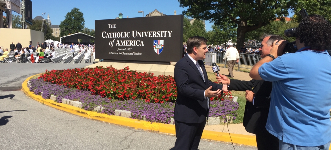 A news reporter films his report in from of a sign welcoming visitors to the CUA campus.